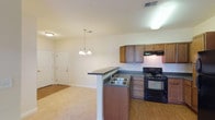 This single-floor unit at The Reserve presents a practical and well-organized layout designed for comfortable everyday living. The kitchen serves as a functional hub, outfitted with warm medium-toned wood cabinetry offering ample upper and lower storage, dark-finish appliances including a range and full-size refrigerator, dark countertops, and a double-basin sink set into a peninsula. The peninsula extends into the adjoining living and dining area, creating an open, connected feel. An elegant chandelier pendant light anchors the dining space, while the living area benefits from natural light streaming through French doors that open to an outdoor patio, providing a seamless indoor-outdoor connection. Flooring transitions thoughtfully between wood-look surfaces in the kitchen and soft carpeting throughout the living and sleeping areas. The bedroom is a well-proportioned, quiet retreat featuring a ceiling fan with light fixture, a broad window admitting natural light, and a tray ceiling detail that adds architectural interest. The bathroom is cleanly appointed with a single-sink vanity topped with a white countertop, a large mirror with overhead lighting, and a full-size soaking tub and shower combination with chrome fixtures.