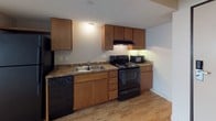 This 1-bedroom, 1-bathroom unit is designed with practicality and comfort in mind across a single-floor layout. The living area features light wood-style flooring, neutral white walls, and a large window fitted with horizontal blinds that allows ample natural light to fill the space. A wall-mounted HVAC unit provides climate control directly in the living area.

The kitchen is efficiently arranged with warm wood cabinetry — both upper and lower — paired with dark granite-toned countertops. Black appliances, including an electric range, dishwasher, and microwave, are integrated seamlessly into the cabinetry, and an overhead range hood adds functional ventilation.

The bedroom is carpeted in a neutral tone and features its own large window with blinds, a wall-mounted HVAC unit, and direct access to the bathroom. The full bathroom includes light tile flooring, a bathtub with surround, a vanity with wood cabinetry, a large mirror, and a grab bar for added accessibility. The textured ceilings are consistent throughout the unit, contributing to a cohesive and understated aesthetic. The overall layout is compact yet well-organized, making efficient use of the available space.