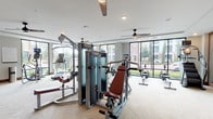 This single-floor fitness center is thoughtfully designed to accommodate a comprehensive range of workout routines. The space is divided into distinct functional zones, each dedicated to a specific training style. The cardio area is highlighted by a curved, glass-walled section lined with floor-to-ceiling windows, filling the room with natural light and offering open outdoor views. This zone houses multiple treadmills, elliptical trainers, a stationary bike, and a rowing machine, all equipped with individual digital displays. The strength training area features a multi-station cable machine along with additional selectorized equipment, providing a full suite of resistance training options. A dedicated free weights zone offers a complete dumbbell rack, adjustable benches, and wall-mounted storage for medicine balls and accessories. A standout feature is the TRX Training Zone, a designated functional fitness area with suspension trainers and a barn-style sliding door that adds a warm, rustic accent to the otherwise modern interior. Throughout the space, herringbone-patterned carpeting, ceiling fans, and recessed lighting contribute to a clean and comfortable atmosphere. Wide corridors connect each training zone, ensuring easy navigation and an open, uncluttered feel across the entire facility.