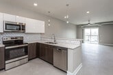 This contemporary single-floor unit offers a thoughtfully designed open-concept layout that seamlessly connects the kitchen and living areas. The kitchen is a standout feature, showcasing dark espresso lower cabinetry paired with high-gloss white upper cabinets, light quartz countertops, a geometric-patterned tile backsplash, and a full suite of stainless steel appliances including a French door refrigerator, electric range, built-in microwave, and dishwasher. Sleek pendant lighting hangs above the peninsula, adding a modern accent. The living area is bright and open, finished with neutral carpet and recessed lighting, with a ceiling fan overhead. A dedicated entryway includes a built-in bench and coat hooks, providing a functional drop zone just inside the front door. The bathroom offers a single-sink vanity with dark wood cabinetry and quartz countertop, alongside a full soaking tub and shower combination. The bedroom connects to a generously sized walk-in closet with wire shelving on both sides. A sliding glass door leads to a private balcony that brings in abundant natural light and open cityscape views. Exposed ductwork in select areas adds an industrial-modern touch to the overall aesthetic.