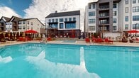 Located at 1801 E 121st Ave in Northglenn, Colorado, this resort-style pool area is a standout amenity designed for relaxation and outdoor living. The centerpiece is a generously sized, crystal-clear swimming pool with a separate spa, both set within a spacious, well-maintained concrete sundeck. Surrounding the pool, an abundance of lounge chairs in two distinct styles — bold red-framed chairs with matching cushions and sleek white-framed chairs with navy cushions — provide ample seating for sunbathing and unwinding. Striped and solid red umbrellas are positioned throughout the deck, offering shaded retreats on sunny Colorado days. A large outdoor screen is mounted near the lounge area, adding an entertainment element to the space. The pool area is enclosed by a modern metal-framed fence with lush greenery and ornamental plantings that add color and privacy. A distinctive red barn-style clubhouse structure anchors one end of the pool deck, complemented by a wooden pergola and additional seating areas. The backdrop features the contemporary multi-story residential buildings of the community, which frame the outdoor space with architectural interest. Open skies above offer expansive views, reinforcing the resort-like atmosphere of this thoughtfully designed outdoor amenity.