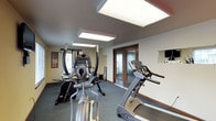 This fitness room is designed to provide a functional and comfortable workout environment. The space is illuminated by large, recessed panel lights mounted in the ceiling, ensuring bright, even lighting throughout the area — ideal for exercising at any time of day. The room features a carpet flooring and neutral-toned walls with warm wood baseboards, creating a clean and practical atmosphere.

The fitness area is equipped with a variety of exercise machines, including a treadmill, an elliptical cross-trainer, and a multi-function strength training machine, offering users a well-rounded workout experience. A wall-mounted television is positioned for convenient viewing during workouts. Motivational fitness posters adorn the walls, adding energy to the space.

Double doors with frosted glass panels provide a stylish entryway into the room, allowing natural light to filter through while maintaining privacy. The overall layout is thoughtfully arranged to maximize usable floor space, ensuring ease of movement between equipment. This fitness room serves as a dedicated space for health and wellness, combining practical amenities with a welcoming and well-maintained environment.