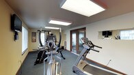 This fitness room is designed to provide a functional and comfortable workout environment. The space is illuminated by large, recessed panel lights mounted in the ceiling, ensuring bright, even lighting throughout the area — ideal for exercising at any time of day. The room features a carpet flooring and neutral-toned walls with warm wood baseboards, creating a clean and practical atmosphere.

The fitness area is equipped with a variety of exercise machines, including a treadmill, an elliptical cross-trainer, and a multi-function strength training machine, offering users a well-rounded workout experience. A wall-mounted television is positioned for convenient viewing during workouts. Motivational fitness posters adorn the walls, adding energy to the space.

Double doors with frosted glass panels provide a stylish entryway into the room, allowing natural light to filter through while maintaining privacy. The overall layout is thoughtfully arranged to maximize usable floor space, ensuring ease of movement between equipment. This fitness room serves as a dedicated space for health and wellness, combining practical amenities with a welcoming and well-maintained environment.