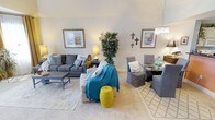 This single-floor residence offers a thoughtfully designed open-concept layout that seamlessly connects the living and dining areas, creating a spacious and cohesive environment. The living area is anchored by a plush gray sofa ensemble accented with decorative pillows, a woven area rug, and a classic coffee table, all contributing to a warm and inviting atmosphere. Pops of color — including a teal throw blanket and a vibrant yellow garden stool — add personality and visual interest to the neutral palette. A tall decorative indoor tree brings a touch of natural greenery to the space, enhancing its lively character. The dining area transitions naturally from the living space, featuring a glass-top table surrounded by upholstered gray chairs and illuminated by an elegant drum pendant light. The walls are adorned with framed artwork and decorative wall accents, adding charm and a curated feel throughout. Soft, light-colored carpeting runs throughout the space, contributing to a sense of warmth and comfort underfoot. The high ceilings amplify the sense of openness, making the entire living area feel airy and expansive. Overall, the space balances classic design elements with contemporary accents for a polished, livable aesthetic.