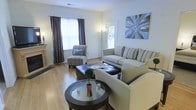 This single-floor space centers around a well-appointed living area that exudes a warm and inviting ambiance. The room features smooth hardwood flooring that extends throughout, contributing to a clean and cohesive aesthetic. Natural light filters in through two windows dressed with elegant purple drape curtains, brightening the neutral white walls and creating an airy feel.

The living area is thoughtfully arranged with a generous sofa and a coordinating accent chair, both upholstered in neutral tones with striped throw pillows that add subtle texture. A dark leather ottoman serves as a functional centerpiece, complemented by a round glass-top coffee table that adds a refined touch. A decorative botanical centerpiece on the table brings a natural element to the space.

Wall décor includes a striking blue and grey abstract artwork that draws the eye and ties together the room's cool color palette. A floor lamp in the corner provides additional ambient lighting. The overall design balances comfort and style, making the living area a versatile and welcoming environment suited for both relaxing and entertaining.