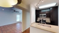 This single-story space offers an open and functional layout connecting the kitchen and living area. The kitchen is well-appointed with dark espresso cabinetry that contrasts sharply against white walls, creating a bold visual dynamic. Stainless steel appliances, including an electric range and built-in microwave, provide a modern, contemporary feel. A peninsula with a light-toned granite or stone countertop and a double-basin stainless steel sink separates the kitchen from the rest of the space, offering both prep functionality and a casual gathering spot. Track lighting overhead ensures the kitchen is well-lit and practical. Warm terracotta tile flooring runs consistently throughout, adding an earthy, inviting quality to the interior. The adjacent living area is visible through a wide archway, where natural light filters in through a horizontal-blind-covered window. A ceiling fan in the living area adds to the comfort and airflow of the space. The overall ambiance is clean and practical, blending modern kitchen finishes with warm, classic flooring for a balanced, livable aesthetic.