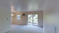 This single-floor space offers a clean, straightforward layout centered around a bright and open living area. The room features wall-to-wall carpeting that adds a soft, neutral base throughout. Natural light flows generously into the space through a sliding glass door that opens directly onto a private balcony, where mature trees create a lush, green backdrop that adds a sense of tranquility and connection to the outdoors. A secondary window on the adjacent wall further enhances the natural brightness of the room. A wall-mounted air conditioning unit provides climate control, ensuring year-round comfort. The smooth, flat ceiling and neutral-toned walls create a simple, understated aesthetic that offers flexibility for a variety of interior styles. The open floor plan allows for versatile furniture arrangements, making it well-suited for use as a living room or multi-purpose area. Overall, the space presents a calm and functional environment, with its standout feature being the seamless indoor-outdoor connection through the balcony access and the natural greenery visible from within.
