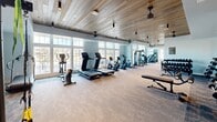 Located in Vestavia Hills, Alabama, this fitness center offers a thoughtfully designed, single-floor layout divided into two distinct workout zones, each tailored to a different style of training. The main workout area features a striking wood-plank ceiling with pendant and track lighting, ceiling fans, and soft blue-gray walls that create an energizing yet comfortable atmosphere. Large multi-pane windows line the walls, flooding the space with natural light and offering views of the surrounding neighborhood. This zone is equipped with a full row of cardio machines — including treadmills, ellipticals, and stationary bikes — as well as cable machines, a dumbbell rack, weight benches, and wall-mounted mirrors that enhance the sense of openness. A built-in wood shelving unit neatly displays yoga blocks, foam rollers, and wellness accessories. The second zone features bright white walls, a tray ceiling with a ceiling fan, and expansive windows on multiple sides that bathe the room in daylight. This area is suited for functional and flexible training, accommodating stability balls, a rowing machine, a pull-up/dip station, and open floor space for stretching or group activity. Plush carpet tile flooring runs throughout both areas, contributing to a polished, community-facility aesthetic.