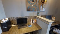 This single-floor space is thoughtfully designed to balance professionalism with a warm, welcoming ambiance. The reception area serves as the focal point, featuring a granite-topped counter with rich, earthy tones that lend an air of quality and sophistication. A mosaic tile backsplash with small, earth-toned tiles adds a decorative texture that enhances the visual depth of the space. The reception desk is equipped with a desktop computer workstation, making it fully functional for administrative use, and is complemented by informational brochures neatly displayed for visitors. The walls are painted in a soft, neutral gray-blue tone that promotes a calm and relaxing environment. Decorative wall art, featuring bold teal and brown floral motifs on a rustic wood background, adds a contemporary artistic touch to the space. A decorative vase with teal and brown tones sits on a ledge, reinforcing the cohesive color palette throughout. Natural light filters in through a nearby window, contributing to the bright and airy feel of the space. Overall, the design reflects a polished, detail-oriented aesthetic suitable for a professional service or wellness-oriented environment.
