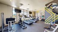 This fitness center offers a comprehensive workout environment designed to accommodate a wide range of exercise routines. The space is organized across a single level and benefits from an abundance of natural light, courtesy of expansive floor-to-ceiling windows and glass doors that frame views of the surrounding outdoor areas.

The cardio zone is well-appointed with multiple treadmills, elliptical machines, and a stair climber, all arranged along the windowed walls to take advantage of the outdoor scenery. Wall-mounted flat-screen televisions are positioned throughout for entertainment during workouts. The strength training area features a cable machine, free weights, medicine balls, stability balls, resistance equipment, and a dedicated bench press station, providing a thorough selection of options for resistance training.

A striking hand-drawn-style mural serves as a bold focal point along one wall, adding an artistic and energetic character to the space. Mirrors line portions of the walls, enhancing the sense of openness. The flooring throughout is durable rubber gym matting, practical and well-suited to the demands of the space.

A small utility corridor provides dual water fountains and restroom access, adding everyday convenience for users. The overall ambiance is bright, modern, and motivating, combining functional fitness infrastructure with thoughtful design elements.