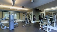 This fitness center is thoughtfully designed to accommodate a well-rounded strength and conditioning routine. The space features dark rubber flooring, which provides both durability and cushioning underfoot, making it well-suited for heavy equipment and high-impact exercise. The walls are painted in a neutral dark tone, lending the room a focused, gym-like atmosphere. Large mirrors line the walls, enhancing the sense of space and allowing users to monitor their form during workouts. The ceiling incorporates recessed lighting alongside a ceiling fan, ensuring the space remains well-lit and well-ventilated during use. Among the notable equipment, a multi-station cable and weight machine serves as the centerpiece, supplemented by a dedicated bench press setup and a free weight rack with dumbbells. Additional strength machines are positioned along the perimeter for a variety of targeted exercises. Large windows allow natural light to filter into the space, providing a pleasant connection to the exterior. Overall, the fitness center is well-organized and equipped to support users at various fitness levels.