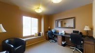 This single-floor office space is designed for functionality and comfort, offering a thoughtfully arranged workspace with several notable features. The room is finished with warm, golden-tan walls that create an inviting atmosphere, complemented by neutral carpeted flooring throughout.

A built-in wooden workstation lines one wall, providing generous desk space equipped with dual computer setups, including monitors, a printer, and ergonomic office chairs. The cabinetry beneath the desk offers ample storage with wooden drawer and cabinet units in a rich, medium-toned finish. A large framed mirror mounted above the workstation adds depth to the room and enhances the sense of space.

Natural light enters through a sizable window fitted with horizontal blinds, beneath which sits a cushioned window bench — a practical and comfortable addition that doubles as extra seating or a relaxation spot. Accent pillows add a touch of décor to this feature.

Overhead, flush-mount ceiling light fixtures provide consistent, even illumination throughout the room. A small table lamp on the desk adds warm, task-specific lighting. The overall ambiance is professional yet comfortable, making this space well-suited for a dedicated home or shared office environment.