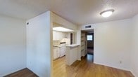 This single-floor residence offers a thoughtfully arranged layout that balances functionality with an open, welcoming atmosphere. The interior is anchored by warm hardwood flooring that flows seamlessly throughout the main living areas, creating a sense of continuity and warmth.

The kitchen is well-appointed with white cabinetry, a white appliance suite including a range and dishwasher, and a built-in sink positioned beneath under-cabinet lighting. A half-wall peninsula separates the kitchen from the adjoining living space, providing both a casual dining or prep surface and an open sightline that keeps the area feeling connected and bright.

Built-in open shelving near the kitchen adds a practical organizational feature while contributing to the overall character of the space. A hallway leads toward additional rooms, with a glimpse of natural light filtering through a window at the far end, hinting at further living quarters beyond.

The textured ceiling throughout adds subtle visual depth, while flush-mounted lighting and ceiling vents reflect a clean, utilitarian design approach. The white walls and light tones across the space amplify brightness and create a neutral backdrop adaptable to a variety of interior styles. Overall, the space presents a clean, move-in-ready environment suited for efficient and comfortable day-to-day living.