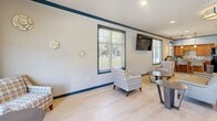 Located at 2001 Amber Leaf Place in Waldorf, Maryland, this single-floor community space combines a fully equipped kitchen and an open living area into one cohesive, functional environment. The interior is finished with light-toned luxury vinyl plank flooring throughout, creating a clean and consistent aesthetic across the entire space. Crisp light gray walls are accented by bold navy blue crown molding and baseboards, lending a polished, distinctive character to the design.

The living area is generously proportioned, furnished with comfortable seating arrangements including patterned accent chairs, sofas, and coffee tables. Multiple large windows fitted with horizontal blinds line the walls, flooding the room with natural light and offering views of the surrounding outdoor greenery. A wall-mounted flat-screen television serves as a central focal point for the lounge area.

The kitchen features warm-toned wood cabinetry, granite countertops with a subway tile backsplash, and stainless steel appliances including a full-size refrigerator, microwave, and sink. Pendant lighting illuminates a kitchen island with bar-height seating. A dual water fountain station and a bulletin board area provide additional practical amenities. The entryway is anchored by a set of glass double doors, welcoming visitors with an open and airy foyer.