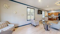 Located at 2001 Amber Leaf Place in Waldorf, Maryland, this single-floor community space combines a fully equipped kitchen and an open living area into one cohesive, functional environment. The interior is finished with light-toned luxury vinyl plank flooring throughout, creating a clean and consistent aesthetic across the entire space. Crisp light gray walls are accented by bold navy blue crown molding and baseboards, lending a polished, distinctive character to the design.

The living area is generously proportioned, furnished with comfortable seating arrangements including patterned accent chairs, sofas, and coffee tables. Multiple large windows fitted with horizontal blinds line the walls, flooding the room with natural light and offering views of the surrounding outdoor greenery. A wall-mounted flat-screen television serves as a central focal point for the lounge area.

The kitchen features warm-toned wood cabinetry, granite countertops with a subway tile backsplash, and stainless steel appliances including a full-size refrigerator, microwave, and sink. Pendant lighting illuminates a kitchen island with bar-height seating. A dual water fountain station and a bulletin board area provide additional practical amenities. The entryway is anchored by a set of glass double doors, welcoming visitors with an open and airy foyer.