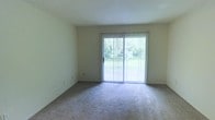 This single-floor residential space offers a clean, functional layout designed for comfortable everyday living. The living area is a defining feature of the space, finished with wall-to-wall carpeting that adds warmth underfoot and contributes to a quiet, cozy atmosphere. Neutral-toned walls keep the interior feeling open and adaptable to a variety of furnishing styles.

A standout element of the living area is the full-width sliding glass door, dressed with vertical blinds that allow for flexible light control. The door frames a verdant outdoor view, flooding the room with abundant natural light and creating a seamless visual connection to the surrounding greenery outside. This feature lends the space an airy quality despite its straightforward design.

The textured ceiling complements the understated aesthetic, maintaining a consistent and uncluttered look throughout. The overall ambiance of the space is calm and neutral, providing a blank canvas well-suited for personalization. With its emphasis on natural light, outdoor connectivity, and practical finishes, this space presents a livable and approachable environment for its occupants.