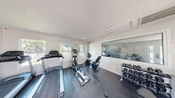 This single-floor community amenity facility comprises two distinct functional spaces: a fitness room and a laundry room, together offering residents convenient on-site services.

The fitness room features dark, durable flooring with protective rubber mats beneath the equipment. It is outfitted with multiple cardio machines, including treadmills, an elliptical, and a stationary bike, alongside a weight bench and a tiered dumbbell rack stocked with a full range of free weights. Natural light fills the room through a row of windows along one wall, offering views of surrounding greenery. A large interior window connects the fitness area to an adjacent space, enhancing the open feel. Access is provided through a glass-paneled door leading to an outdoor area.

The laundry room is bright and clean, finished with white tile flooring and recessed ceiling lighting. It is lined with multiple full-size washing machines and dryers along the walls, accompanied by a central stainless steel folding table for resident use. A payment kiosk is available for added convenience, and seating along one wall provides a comfortable area to wait. A window allows natural light to enter the space, and a view of the pool area is visible from within.

Together, these two spaces form a practical and well-maintained amenity suite designed to serve the daily needs of a residential community.