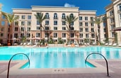Located at 13732 Jamboree Rd in Irvine, California, this outdoor amenity space presents a well-appointed resort-style courtyard enclosed by a multi-story building with a classic, warm-toned stucco facade and large windows. The centerpiece of the space is a generously sized swimming pool with clear turquoise water, complemented by a separate spa featuring decorative tile detailing and dual entry handrails. The pool deck is finished in brick-patterned pavers and is surrounded by an abundance of seating options, including rows of white and neutral-toned chaise lounges positioned on both hardscape and synthetic turf areas. Large cantilever umbrellas in muted rose and terracotta tones provide ample shade throughout the deck. The courtyard is framed by mature palm trees that rise above the roofline, adding a tropical ambiance, while lush, manicured planting beds filled with tropical foliage, flowering shrubs, and ornamental grasses line the perimeter fencing. Wrought iron fencing with vertical slat detailing separates distinct zones within the outdoor space. Amenities include outdoor dining tables, free Wi-Fi access points, and a smoke-free environment. The space is well-lit for evening use, creating an inviting atmosphere day and night.