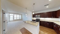 This single-floor space showcases an open-concept layout that seamlessly connects the kitchen and living area. The kitchen is outfitted with dark espresso-toned cabinetry, stainless steel appliances, and a light-colored countertop island that doubles as a prep and gathering space. Pendant lighting hangs above the island, adding a modern touch, while recessed ceiling lights provide even illumination throughout the kitchen. The flooring in the kitchen is a rich dark hardwood, creating a striking contrast with the neutral-toned carpet that extends across the generous living area. The living space is bright and open, with clean white walls and standard-height ceilings that contribute to an airy feel. A hallway visible from the main living area leads to additional rooms, including a bathroom that features a toilet and what appears to be shelving or closet storage. The overall aesthetic is contemporary and neutral, with a practical, functional layout that prioritizes open flow between the main living and kitchen zones. The space presents a clean, move-in-ready appearance throughout.
