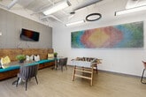 Located at 349 Decatur St SE in Atlanta's Grant Park neighborhood, this well-appointed community lounge is designed to foster social interaction and leisure. The single-floor space features an open layout with light wood-look flooring, exposed ductwork, and high ceilings that lend it an airy, industrial-modern character. Stylish circular and linear pendant lighting illuminates the room effectively throughout.

The lounge is anchored by a full-size foosball table positioned beneath a striking, large-scale colorful wall-mounted artwork that serves as the room's visual centerpiece. A built-in tufted bench with teal upholstery and accent pillows runs along one wall, complemented by a wall-mounted flat-screen television above textured wallcovering — creating an inviting nook for casual gathering. Multiple round tables paired with contemporary upholstered chairs are distributed throughout, offering flexible seating arrangements.

A dedicated coffee and refreshment bar is built into warm wood cabinetry, providing a functional amenity for residents or guests. Floor-to-ceiling glass doors and windows along one wall flood the space with natural light and provide views of the surrounding outdoor courtyard and greenery, creating a seamless connection to the exterior. The overall ambiance is modern, energetic, and welcoming.