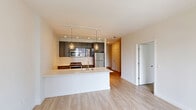Located in Chicago's vibrant River North neighborhood, this modern one-bedroom, one-bathroom residence offers a thoughtfully designed open-concept layout with contemporary finishes throughout. Light-toned hardwood flooring flows through the main living areas, anchored by a sleek kitchen that serves as the centerpiece of the space. The kitchen features high-gloss gray cabinetry, stainless steel appliances including a gas range, microwave, and refrigerator, white quartz countertops, and a peninsula island with pendant lighting overhead — ideal for both cooking and entertaining. The living area is bright and open, with clean white walls and a neutral palette. A hallway leads to the spacious bedroom, which is carpeted and framed by large floor-to-ceiling windows offering sweeping views of the Chicago skyline. Exposed concrete columns add an industrial architectural touch to the bedroom's modern aesthetic. The bathroom is finished with classic white subway tile surrounding a soaking tub and shower combo, complemented by brushed nickel fixtures. A stacked washer and dryer unit is conveniently tucked into a dedicated closet along the hallway, adding practical in-unit laundry. The overall ambiance is clean, bright, and urban.
