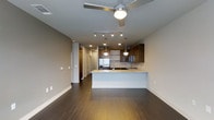 This contemporary single-floor residence in Dallas offers a thoughtfully designed open-concept layout that seamlessly blends living and kitchen spaces. Rich, dark hardwood flooring extends throughout, grounding the space with warmth and sophistication. The kitchen is a standout feature, showcasing rich wood cabinetry, a light-toned stone countertop island with an undermount sink, and stainless steel appliances including a French door refrigerator. Pendant lighting with globe fixtures hangs elegantly over the island, complemented by recessed lighting and a track lighting system that together provide layered illumination. The living area is topped with a modern ceiling fan, enhancing both comfort and style. Neutral taupe-toned walls create a calm, versatile backdrop throughout the space. A hallway visible from the main area leads to additional rooms, lined with what appears to be closet or storage access, adding to the functional layout. The overall ambiance is clean, modern, and urban — well-suited for those seeking a refined living environment in the heart of Dallas.