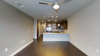 This contemporary single-floor residence in Dallas offers a thoughtfully designed open-concept layout that seamlessly blends living and kitchen spaces. Rich, dark hardwood flooring extends throughout, grounding the space with warmth and sophistication. The kitchen is a standout feature, showcasing rich wood cabinetry, a light-toned stone countertop island with an undermount sink, and stainless steel appliances including a French door refrigerator. Pendant lighting with globe fixtures hangs elegantly over the island, complemented by recessed lighting and a track lighting system that together provide layered illumination. The living area is topped with a modern ceiling fan, enhancing both comfort and style. Neutral taupe-toned walls create a calm, versatile backdrop throughout the space. A hallway visible from the main area leads to additional rooms, lined with what appears to be closet or storage access, adding to the functional layout. The overall ambiance is clean, modern, and urban — well-suited for those seeking a refined living environment in the heart of Dallas.