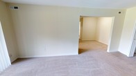 This single-floor space presents a straightforward and functional layout designed to maximize usability. The interior is characterized by soft, neutral-toned walls that create a calm and welcoming atmosphere throughout. Plush wall-to-wall carpeting in a light beige tone runs seamlessly across the living areas, adding warmth and comfort underfoot. The main living area is open and generously proportioned, flowing naturally into an adjoining room through a wide, arched passageway — a thoughtful architectural detail that enhances the sense of connectivity and space. The adjoining room mirrors the same neutral palette and carpeted flooring, maintaining visual consistency across the layout. Natural light filters in through windows visible in the secondary room, brightening the interior and contributing to the overall airy feel. Clean white baseboards provide a crisp contrast against the carpeting, adding a subtle finishing touch to the interior. The space is currently vacant, offering a blank canvas that allows for flexible furniture arrangements and personalization. Overall, the design is simple, cohesive, and practical, suited for a variety of uses and lifestyles.