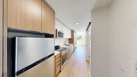 Unit 504 at 1140 NW Market St in Seattle's Ballard neighborhood is a thoughtfully designed studio that maximizes every inch of its compact footprint. Light oak hardwood flooring runs throughout, lending warmth and continuity to the open layout.

The galley-style kitchen is a standout feature, outfitted with a combination of white upper cabinetry and natural wood-toned lower cabinets, a white tile backsplash, stainless steel appliances including a range, built-in microwave, refrigerator, and dishwasher, and a black matte sink faucet. A large window at the end of the kitchen corridor fills the space with natural light and frames a lush green treetop view.

The main living area is open and airy, anchored by a floor-to-ceiling built-in wardrobe in a warm wood finish, offering generous storage. A large window along the exterior wall allows ample natural light to pour in. A bold blue entry door adds a striking design accent upon arrival.

The bathroom is clean and modern, featuring wood-toned upper cabinetry, a tiled walk-in shower with matte black fixtures, and wood-look flooring that maintains design cohesion with the rest of the unit. In-unit laundry is also present near the entry. The overall ambiance is bright, minimal, and efficiently designed.