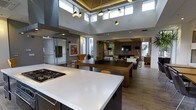 This thoughtfully designed single-floor space showcases an open-concept layout that seamlessly connects the kitchen, dining, and living areas into one expansive, flowing environment. The kitchen anchors the space with a large island featuring a sleek white countertop and a built-in gas cooktop, offering both functionality and style. A dark wood dining table sits centrally, complemented by warm-toned chairs that add a touch of character.

The living area at the far end is anchored by a wall-mounted television set against a striking slatted wood accent wall, with a fireplace below creating a warm and inviting focal point. Comfortable seating and soft textiles contribute to the relaxed ambiance of this zone.

Soaring ceilings with clerestory windows flood the interior with natural light, enhancing the sense of openness and airiness throughout. Recessed lighting and a pair of distinctive pendant light fixtures provide layered illumination. The warm wood-toned flooring runs continuously across the space, tying all areas together cohesively.

Additional enclosed spaces are visible along the perimeter, suggesting private rooms or utility areas. Large windows and glass elements throughout connect the interior to the outdoors, reinforcing a bright, modern aesthetic that balances openness with comfort.