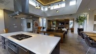 This thoughtfully designed single-floor space showcases an open-concept layout that seamlessly connects the kitchen, dining, and living areas into one expansive, flowing environment. The kitchen anchors the space with a large island featuring a sleek white countertop and a built-in gas cooktop, offering both functionality and style. A dark wood dining table sits centrally, complemented by warm-toned chairs that add a touch of character.

The living area at the far end is anchored by a wall-mounted television set against a striking slatted wood accent wall, with a fireplace below creating a warm and inviting focal point. Comfortable seating and soft textiles contribute to the relaxed ambiance of this zone.

Soaring ceilings with clerestory windows flood the interior with natural light, enhancing the sense of openness and airiness throughout. Recessed lighting and a pair of distinctive pendant light fixtures provide layered illumination. The warm wood-toned flooring runs continuously across the space, tying all areas together cohesively.

Additional enclosed spaces are visible along the perimeter, suggesting private rooms or utility areas. Large windows and glass elements throughout connect the interior to the outdoors, reinforcing a bright, modern aesthetic that balances openness with comfort.