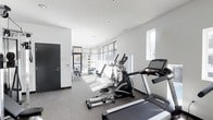 This fitness center offers a bright, contemporary workout environment designed with functionality and comfort in mind. The space is finished with neutral grey carpeted flooring and crisp white walls, creating a clean, modern aesthetic throughout. Natural light floods the room through large floor-to-ceiling windows fitted with horizontal blinds, allowing occupants to control brightness while maintaining an open, airy atmosphere. Additional high-placed windows further enhance ventilation and daylight penetration.

The cardio section is well-equipped, featuring treadmills and an elliptical trainer from recognized brands, positioned to take advantage of the natural light from the windows. Toward the rear of the space, a dedicated strength and flexibility area is visible, outfitted with weight benches, resistance equipment, and medicine balls mounted on the wall for convenient access. Recessed ceiling lighting supplements natural light, ensuring the space remains well-illuminated at all times.

Access is provided through a solid dark-framed door, contributing a bold contrast to the otherwise light interior palette. The overall layout is thoughtfully organized, providing ample circulation space between equipment for safe and comfortable use.
