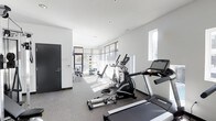 This fitness center offers a bright, contemporary workout environment designed with functionality and comfort in mind. The space is finished with neutral grey carpeted flooring and crisp white walls, creating a clean, modern aesthetic throughout. Natural light floods the room through large floor-to-ceiling windows fitted with horizontal blinds, allowing occupants to control brightness while maintaining an open, airy atmosphere. Additional high-placed windows further enhance ventilation and daylight penetration.

The cardio section is well-equipped, featuring treadmills and an elliptical trainer from recognized brands, positioned to take advantage of the natural light from the windows. Toward the rear of the space, a dedicated strength and flexibility area is visible, outfitted with weight benches, resistance equipment, and medicine balls mounted on the wall for convenient access. Recessed ceiling lighting supplements natural light, ensuring the space remains well-illuminated at all times.

Access is provided through a solid dark-framed door, contributing a bold contrast to the otherwise light interior palette. The overall layout is thoughtfully organized, providing ample circulation space between equipment for safe and comfortable use.