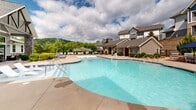 Located at 105 Exchange Circle in Asheville, North Carolina, this single-level outdoor amenity space offers a well-appointed recreational environment designed for leisure and community enjoyment. At its centerpiece is a generously sized, freeform swimming pool with crystal-clear water, featuring built-in water jets, entry handrails, and a shallow wading area — all set against a backdrop of rolling, tree-covered hillsides that are characteristic of the Blue Ridge region.

Adjacent to the pool, a covered outdoor lounge pavilion provides a shaded gathering space furnished with comfortable sectional seating and accent chairs, accented by stone-clad columns and a ceiling fan for year-round usability. The pool deck wraps around the water with ample space for lounge chairs and thatched-roof cabana-style shade structures, creating a resort-like atmosphere.

A gated entry leads into the pool area, reinforcing both security and a sense of arrival. To the side of the main pool complex, a serene green lawn area features a freestanding hammock framed by manicured evergreen trees and Adirondack seating — an inviting spot for quiet relaxation. Stone-accented landscaping with native plantings and river rock borders tie the entire outdoor space together, complementing the multi-story residential buildings that rise in the background.