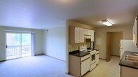 This single-floor space presents an open-concept layout that connects the kitchen and living area, creating a functional and cohesive environment. The kitchen is equipped with white cabinetry featuring wood-trimmed accents, dark laminate countertops, and white appliances including an electric range with oven. An overhead exhaust hood is mounted above the cooktop, and ample upper cabinet storage lines the wall. The kitchen transitions seamlessly into the living area, which is finished with wall-to-wall carpeting and neutral-toned walls, contributing to a clean and understated aesthetic. A vertical blind-covered window or sliding door at the far end of the living area allows natural light to filter into the space. The flooring in the kitchen and adjacent entry areas consists of light-toned hard-surface flooring, providing a practical contrast to the carpeted living zone. An electrical panel or utility access box is visible along one wall near the kitchen. The overall layout is straightforward and efficiently organized, making good use of the available floor plan across this single level.