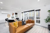 This single-floor space showcases a thoughtfully designed open-plan layout that balances modern aesthetics with inviting, character-rich details. The main lounge area is anchored by a tufted tan leather sectional sofa, complemented by sleek black leather chairs arranged around a live-edge wood coffee table. Large sliding glass doors flood the space with natural light and open onto a private balcony with views of the surrounding urban streetscape. Recessed lighting and clean white walls throughout maintain a bright, airy atmosphere.

A dedicated media area features a wall-mounted large-format television above a minimalist credenza, creating a functional entertainment zone. Adjacent to the main lounge, a secondary seating nook is adorned with eclectic Pacific Northwest–themed wall décor, including pennants, a PNW sign, and a decorative deer mount, lending the space a distinctive regional personality.

The kitchenette stands out as a visual centerpiece, showcasing a bold multicolored geometric tile backsplash in rich jewel tones. It is equipped with dark cabinetry, a dark stone countertop, an undermount sink, a built-in microwave, and a beverage refrigerator, making it fully functional for entertaining. Light-toned tile flooring runs throughout, tying the various zones together cohesively.
