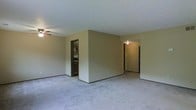 This single-floor space presents an open and functional layout highlighted by neutral beige walls and wall-to-wall plush carpeting throughout. The main living area is spacious, offering generous room for furniture arrangement, and is equipped with a ceiling fan to promote air circulation. A central dividing wall creates a natural separation within the open floor plan while maintaining a sense of connectivity between areas. Two distinct doorways are visible from the main living space — one leading to what appears to be a kitchen or utility area featuring dark cabinetry and appliances, and another corridor-style opening that leads further into the unit, likely toward additional rooms. The ceiling fan mounted in the secondary area beyond the dividing wall suggests another functional living zone. The consistent carpeting and neutral color palette throughout the visible areas contribute to a cohesive, understated aesthetic. The overall ambiance is clean and straightforward, offering a blank canvas well-suited for personalization. The layout's open sightlines and multiple access points between rooms enhance the sense of flow and accessibility throughout the space.