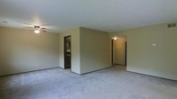 This single-floor space presents an open and functional layout highlighted by neutral beige walls and wall-to-wall plush carpeting throughout. The main living area is spacious, offering generous room for furniture arrangement, and is equipped with a ceiling fan to promote air circulation. A central dividing wall creates a natural separation within the open floor plan while maintaining a sense of connectivity between areas. Two distinct doorways are visible from the main living space — one leading to what appears to be a kitchen or utility area featuring dark cabinetry and appliances, and another corridor-style opening that leads further into the unit, likely toward additional rooms. The ceiling fan mounted in the secondary area beyond the dividing wall suggests another functional living zone. The consistent carpeting and neutral color palette throughout the visible areas contribute to a cohesive, understated aesthetic. The overall ambiance is clean and straightforward, offering a blank canvas well-suited for personalization. The layout's open sightlines and multiple access points between rooms enhance the sense of flow and accessibility throughout the space.