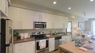 This single-floor residence offers a thoughtfully designed living space centered around a sleek, modern kitchen. The kitchen is outfitted with a full suite of stainless steel appliances, including a freestanding electric range, over-the-range microwave, dishwasher, and refrigerator, all contributing to a cohesive and functional aesthetic. Crisp white cabinetry extends from counter height to the ceiling, maximizing storage while maintaining a clean, contemporary look. Dark countertops provide a striking contrast against the white cabinetry, adding depth and a sense of sophistication to the space. Recessed lighting illuminates the kitchen evenly, enhancing the bright and airy atmosphere. A double-basin sink is positioned near a window, allowing natural light to filter in and brighten the workspace. The open layout creates a natural flow into the adjacent dining area, where a wooden dining table adds warmth and a welcoming touch. Light hardwood-style flooring ties the spaces together seamlessly. Overall, this residence combines practical functionality with modern design elements, creating a comfortable and inviting environment well-suited for everyday living and entertaining.