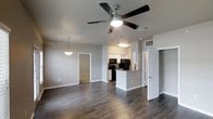 This single-story space showcases an open-concept layout that seamlessly connects the living area to the kitchen, creating a cohesive and functional floor plan. Dark hardwood flooring extends throughout, providing a rich, grounding contrast to the light gray walls and crisp white trim.

The living area is equipped with a modern ceiling fan featuring an integrated light fixture, ensuring both comfort and illumination. A pendant light is visible in the background, adding a subtle decorative touch and helping to define separate zones within the open layout.

The kitchen is positioned at the rear and features white cabinetry, black appliances including a microwave and refrigerator, and a kitchen island with a light-colored countertop that serves as a natural dividing point between the cooking and living spaces. The kitchen is well-lit with overhead lighting that highlights the workspace effectively.

Adjacent to the main living area, a doorway leads to an additional room, while a partially open door on the right reveals a closet with a clothes rod, suggesting a bedroom entry nearby. The overall ambiance is clean, modern, and move-in ready, with a neutral palette that offers broad design flexibility.