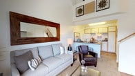 This two-story unit is defined by its dramatic vaulted ceilings, which lend the main living area a remarkably open and airy quality. The living room benefits from this vertical space, with tall walls accommodating a large wood-framed mirror that adds warmth and depth to the room. Neutral carpet and large windows allow natural light to flow freely throughout, creating a bright and welcoming atmosphere.

The kitchen is positioned adjacent to the living area in an open-concept layout, featuring white cabinetry with wood trim, granite-look countertops, a full suite of white appliances including a refrigerator, electric range, and a breakfast bar that provides casual seating and a natural division between the two spaces.

The bathroom is well-appointed with wood-look flooring, a granite-topped vanity, and a full tub-shower combination, offering a clean and functional design.

The upper loft level, accessed via an interior staircase, serves as the bedroom. The space features a sloped, textured ceiling with a skylight that introduces additional natural light. A closet with a hanging rod provides practical storage, and the open railing overlooking the main floor reinforces the loft's connected, spacious character.
