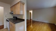 This single-floor unit showcases a practical and straightforward layout that makes efficient use of its space. The kitchen is positioned at the entry of the unit and features light maple wood cabinetry, offering ample storage with both upper and lower cabinet configurations. Dark granite-style countertops provide a contrasting visual element against the warm wood tones of the cabinetry. A gas range is integrated into the kitchen setup, making the space well-equipped for everyday cooking needs.

A structural column separates the kitchen from the main living area, which opens into a generously proportioned room with gleaming hardwood floors that run throughout the unit. The warm honey tones of the flooring lend a sense of continuity and warmth across the space. Neutral white walls throughout keep the interior feeling bright and clean, providing a versatile backdrop.

A hallway visible from the living area leads to additional rooms, suggesting a thoughtful floor plan with clear separation between living and private spaces. The overall ambiance is simple and functional, with classic finishes that offer a solid foundation for personalization.