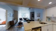 This contemporary single-floor residence showcases a thoughtfully designed open-concept layout that seamlessly connects the kitchen, dining, and living areas. The kitchen is anchored by a generous granite-topped island with a stainless steel sink and gold-toned faucet, complemented by white cabinetry and a stainless steel range. The island serves as a natural focal point, creating an ideal space for both meal preparation and casual entertaining.

The living area features rich dark hardwood flooring throughout, adding warmth and contrast to the light-toned walls. A large picture window floods the space with natural light while offering expansive views of the surrounding urban skyline. The living room is framed by a tray ceiling detail that adds architectural interest and depth to the space.

A dedicated seating nook near the window provides an intimate spot for conversation or relaxation, while built-in shelving along the far wall offers practical display and storage solutions. The overall ambiance is modern and urban, with clean lines, neutral tones, and carefully curated furnishings that emphasize both style and functionality. Track lighting enhances the contemporary aesthetic while providing flexible illumination throughout the open living space.