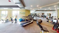 This single-level FitCamp fitness facility is thoughtfully designed to support health and wellness programs. The main exercise floor is generously proportioned, featuring patterned carpet tiles and floor-to-ceiling mirrors that visually expand the space. The area is well-stocked with cardio equipment including treadmills, rowing machines, ellipticals, and stability balls, all arranged to allow comfortable movement throughout the room. Recessed lighting illuminates the space evenly, while large windows along the perimeter bring in natural light and outdoor views, creating an energizing environment.

Adjacent to the exercise floor is a welcoming service station outfitted with warm wood cabinetry, a long countertop, open cubbies stocked with neatly rolled towels, and a wall-mounted display screen — offering a polished, organized point of interaction. A separate open area serves as a consultation and administrative zone, featuring a built-in desk, additional wood cabinetry, seating, and a flat-screen television. This space is bright and airy, with access to the outdoors through glass doors. Together, the zones create a cohesive, professional environment well-suited for guided fitness and rehabilitation programs.