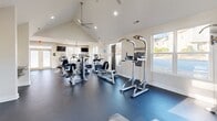 Located at 5855 Old Oak Ridge Rd in Greensboro, NC, this single-floor fitness center is designed to provide a comprehensive and welcoming workout environment. The main exercise room is the heart of the facility, characterized by soaring vaulted ceilings that lend an open, airy feel to the space. Abundant natural light pours in through multiple windows, creating a bright and energizing atmosphere. The room features durable dark flooring throughout and is outfitted with a wide array of equipment, including recumbent bikes, ellipticals, a treadmill, a multi-station cable machine, free weights with a tiered dumbbell rack, and a weight bench. Large mirrors line one wall to support proper form, while two flat-screen TVs and a ceiling fan add to user comfort. A motivational "live fit" wall graphic reinforces the facility's wellness-focused identity. A built-in water fountain station with a bottle-filling option is conveniently located near the entrance. The two bathrooms are thoughtfully designed with accessibility in mind, each featuring ADA-compliant grab bars, warm neutral walls, and wood-look vinyl flooring, ensuring comfort and safety for all users. This facility is a fully functional, purpose-built space ideal for an active community.