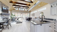 Located at 400 Boulder Pass in Oxford, CT, this single-story community clubhouse offers an impressive blend of rustic charm and modern amenities. The centerpiece of the space is a grand open-concept lounge and social area showcasing dramatic vaulted ceilings with exposed dark wood beams, tongue-and-groove wood ceiling panels, and abundant natural light streaming through clerestory windows and large sliding glass doors that open to an outdoor patio. A floor-to-ceiling stacked stone fireplace with a wood mantel and wall-mounted television anchors the lounge, which is furnished with comfortable seating arrangements and a dining table, creating an inviting atmosphere for gatherings. The full kitchen is outfitted with white shaker-style cabinetry, dark granite countertops, stainless steel appliances, and draft tap fixtures, making it ideal for entertaining. Light-toned hardwood flooring flows throughout the main living and kitchen areas, contributing to a clean, airy aesthetic. A dedicated fitness center features a full complement of cardio machines, a dumbbell rack, kettlebells, resistance equipment, wall-to-wall mirrors, and large windows that fill the room with natural light. The space is thoughtfully designed to serve as a versatile hub for recreation, socializing, and wellness within the community.