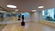 This single-floor fitness studio is thoughtfully designed to support a wide range of training activities. The open floor plan provides ample room for movement, anchored by smooth, light-toned hardwood flooring that lends a clean and welcoming aesthetic throughout the space. A full wall of floor-to-ceiling mirrors lines one side of the studio, enhancing the sense of space while allowing practitioners to monitor their form during workouts. The studio is equipped with a freestanding Everlast punching bag, making it well-suited for boxing and martial arts training, in addition to general fitness use. Overhead, a series of recessed circular light fixtures are evenly distributed across the white ceiling, providing consistent, well-diffused illumination across the entire floor area. Large windows along the perimeter invite generous natural light, creating a bright and energizing atmosphere. A mounted flat-screen display is also visible, offering the ability to follow along with instructional content or virtual classes. The walls are finished in a crisp white, contributing to the studio's clean, modern feel. A private door provides discreet access, adding a functional element to the overall layout. This studio is a well-appointed space for fitness, training, and wellness activities.