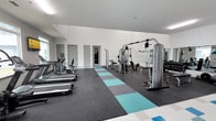 This single-floor fitness center offers a spacious and well-organized workout environment designed to accommodate a variety of exercise routines. The room features high ceilings and bright white walls that create an open, airy atmosphere, enhancing the sense of space throughout. The flooring consists of durable dark rubberized material accented by a central runner of alternating teal, gray, and charcoal interlocking tiles, adding both visual interest and functional cushioning for floor-based exercises. Along one side of the room, a lineup of cardio machines — including treadmills and ellipticals — provides ample options for aerobic training. On the opposite side, a multi-station cable machine and strength training equipment are positioned alongside a large wall mirror, allowing users to monitor their form during workouts. Free weights and benches are also present, rounding out the strength training offerings. Multiple doors along the back wall provide access to additional areas, and recessed lighting keeps the space uniformly well-lit. The overall design is clean, modern, and functional, making it a practical and inviting facility for fitness use.