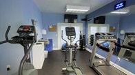 This single-floor fitness center is thoughtfully designed to provide a comprehensive workout environment in a compact, efficient layout. The room features light blue walls that lend a calm, energizing ambiance, complemented by dark rubber flooring ideal for high-impact exercise. Natural light filters in through multiple windows dressed with white blinds, creating a bright and inviting atmosphere. A wall-mounted flat-screen TV is positioned for easy viewing during workouts, adding an entertainment element to training sessions. The space is equipped with a variety of cardio machines, including treadmills and an elliptical trainer, catering to different aerobic exercise preferences. A well-organized free weight rack holds a full range of dumbbells, supporting strength training routines. Additional fitness accessories, such as stability balls and exercise mats, are also available. A large mirror on one wall enhances the sense of space and allows users to monitor their form. A bulletin board and informational materials near the entrance keep users informed. The overall layout is practical and user-friendly, making this fitness center a functional and accessible resource for daily exercise.