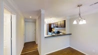 This single-floor, one-bedroom, one-bathroom residence offers a functional and well-designed layout that maximizes the available space. The kitchen is a standout feature, showcasing dark espresso-toned cabinetry that contrasts elegantly with a light, patterned tile backsplash. A breakfast bar-style pass-through counter connects the kitchen to the adjacent living area, creating an open and sociable atmosphere while maintaining a clear sense of purpose for each zone. Stainless steel appliances add a modern, polished touch to the kitchen, and overhead track lighting ensures the workspace is well-illuminated. Crown molding runs along the ceiling throughout the visible areas, adding a refined architectural detail that elevates the overall feel of the interior. Warm hardwood-style flooring extends through the entryway and living areas, providing a cohesive and inviting aesthetic. The entry corridor leads to a solid paneled front door, offering a welcoming and secure point of arrival. Overall, the space presents a clean, neutral palette with thoughtful design details that balance practicality and style.